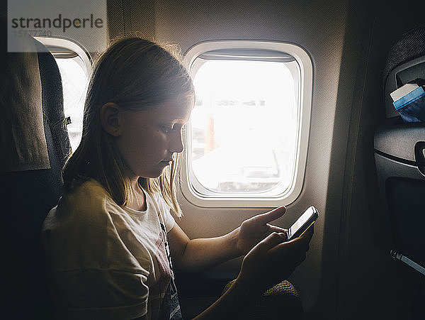 Kaukasisches Mädchen benutzt Mobiltelefon  während sie am Flugzeugfenster sitzt
