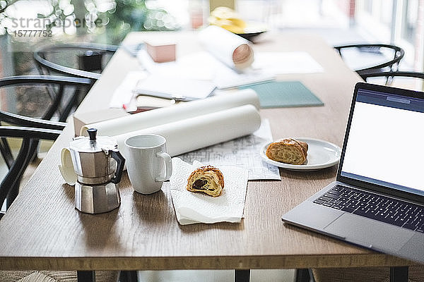 Hochwinkelansicht einer Teekanne mit aufgerollten Dokumenten per Laptop und Croissant auf dem Tisch