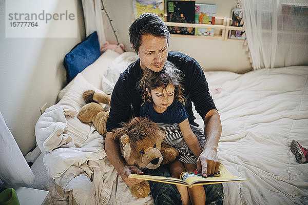 Hochwinkelansicht eines Vaters  der mit seiner Tochter ein Buch liest  während er im Schlafzimmer sitzt