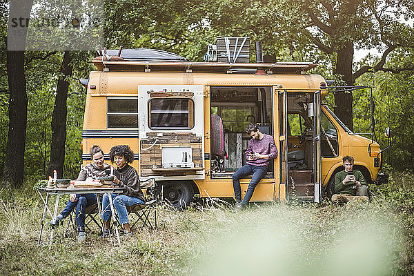 Lächelnde Freundinnen sitzen am Tisch  während Männer beim Camping im Wald Smartphones benutzen