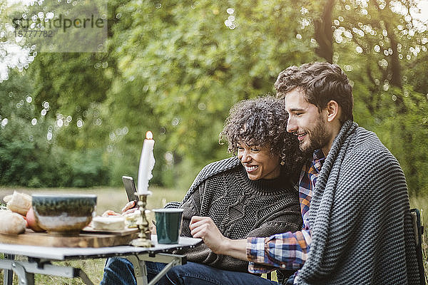Fröhliches junges Paar  das sich während des Zeltens im Wald gemeinsam ein Handy teilt  während es zusammen in eine Decke gehüllt ist