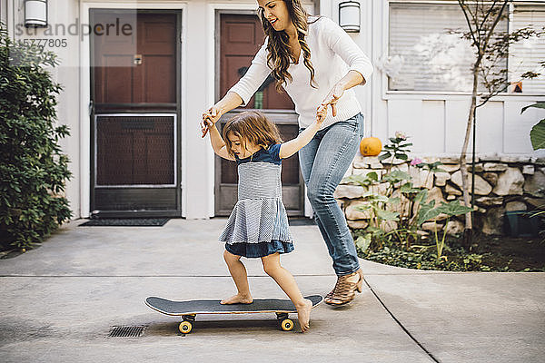 Lächelnde Tochter balanciert über Skateboard mit Hilfe der Mutter auf dem Fußweg