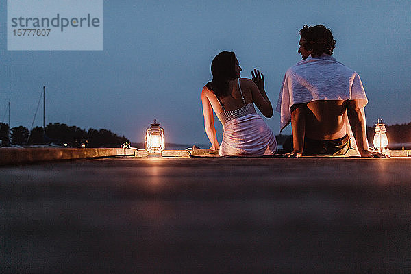 Lächelndes Paar sitzt um eine beleuchtete Laterne am Pier gegen den Himmel