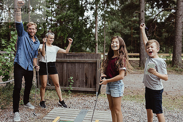 Fröhliche Familie mit erhobenen Armen beim Minigolfspielen im Hinterhof