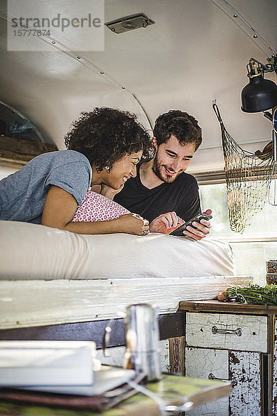 Lächelndes Paar  das sich ein Mobiltelefon teilt  während es im Wohnwagen auf dem Bett liegt