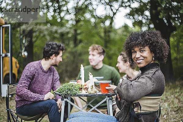 Porträt einer lächelnden Afro-Frau  die beim Camping im Wald mit Freunden am Tisch sitzt