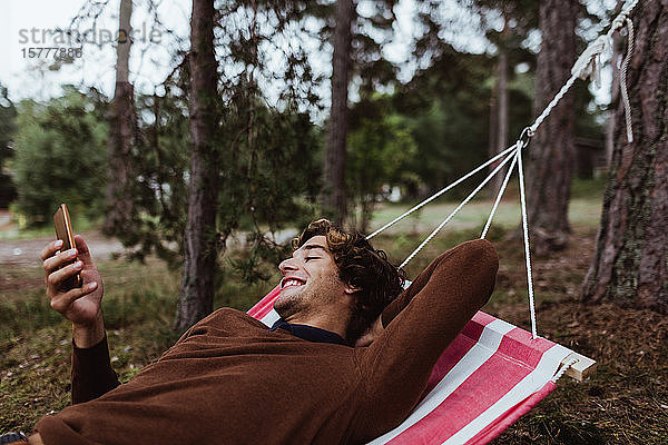 Lächelnder junger Mann benutzt Mobiltelefon  während er sich in Hängematte gegen Bäume entspannt