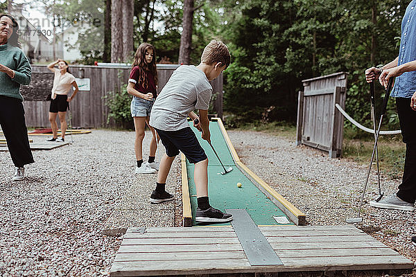 Familie schaut einem Jungen zu  der beim Minigolfen im Garten einen Schuss abgibt