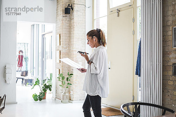 Unternehmerin spricht über ein Smartphone  während sie im Heimbüro ein Dokument betrachtet