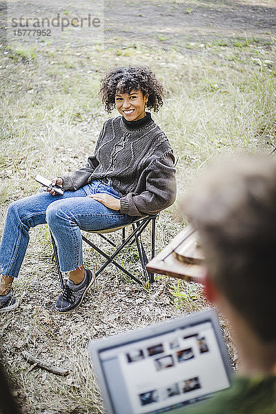 Lächelnde junge Frau sitzt auf einem Stuhl  während sie einen männlichen Freund beim Zelten im Wald ansieht