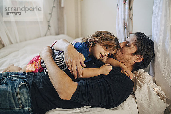 Reifer Vater mit geschlossenen Augen küsst Tochter im Bett liegend