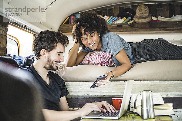 Glückliche junge Frau zeigt einem Mann  der mit einem Laptop im Wohnmobil sitzt  ein Handy
