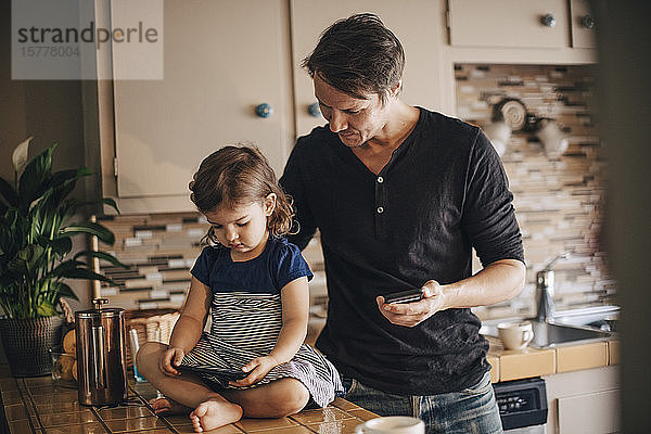 Tochter benutzt Smartphone  während der Vater in häuslicher Küche steht