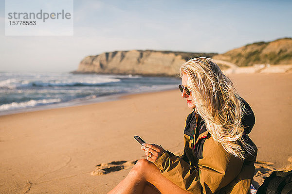 Frau hält Telefon am Strand sitzend