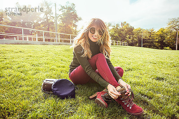 Porträt einer blonden Frau mit Sonnenbrille  die im Gras sitzt und lächelnd in die Kamera schaut
