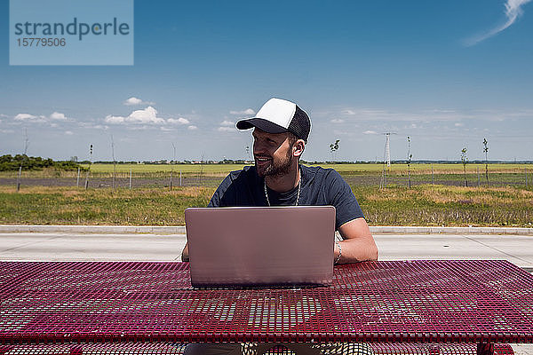 Mann mit Baseballmütze sitzt mit Laptop am Picknicktisch am Straßenrand