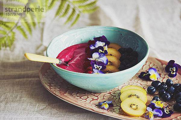 Hochwinkel-Nahaufnahme einer Schale mit rosa Fruchtpüree mit Heidelbeeren und Scheiben von gelben Kiwis  dekoriert mit Stiefmütterchenblättern.