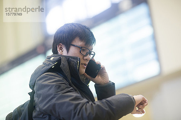 Junger asiatischer Brillenträger steht im Bahnhofsfoyer  schaut auf seine Uhr und benutzt sein Mobiltelefon.