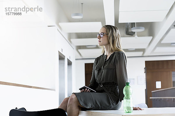 Porträt einer jungen blonden Geschäftsfrau mit Brille  die im Büro auf dem Schreibtisch sitzt.