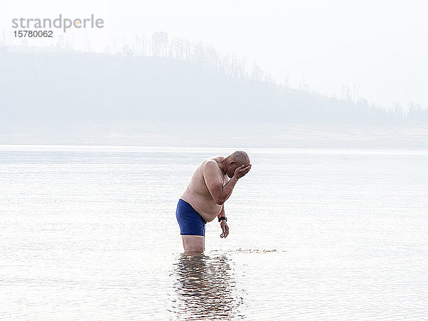 Älterer Mann steht mit dem Oberschenkel tief im Wasser und wäscht sich das Gesicht. Hitze und Rauchschleier in der Luft von Buschfeuern.