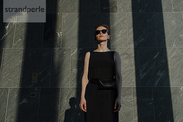 Rothaarige Frau mit Sonnenbrille  vor einer Steinmauer mit Schatten