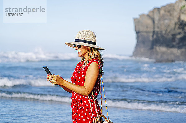 Blonde Frau trägt rotes Kleid und Hut und benutzt Smartphone am Strand