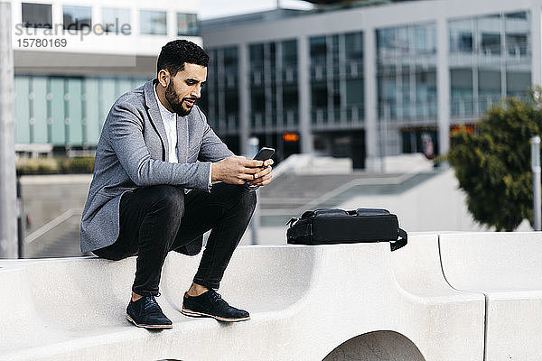 Lässiger junger Geschäftsmann  der auf einer Bank in der Stadt sitzt und sein Handy benutzt
