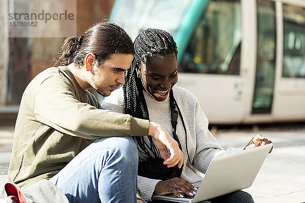 Glückliches junges Paar mit Laptop an der Straßenbahnhaltestelle in der Stadt