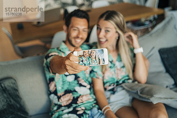 Glückliches Paar sitzt auf der Couch im Wohnzimmer und trägt hawaiianische Hemden und macht einen Selfie