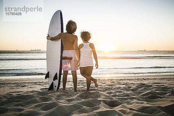 Rückansicht von zwei Jungen mit Surfbrett  die bei Sonnenuntergang am Strand stehen