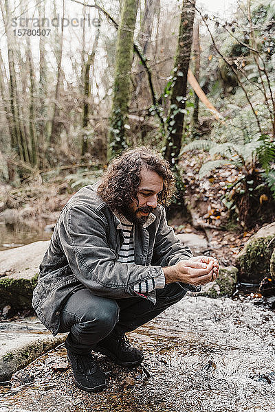 Junger Mann kauert am Bach im Wald und trinkt Wasser aus geschröpften Händen