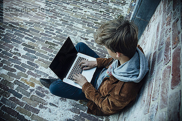 Teenager  der einen Laptop benutzt und auf einem Steinboden in der Stadt sitzt