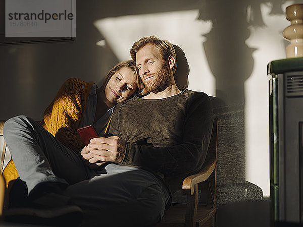 Entspanntes Paar sitzt zu Hause mit Mann auf Bank im Sonnenlicht und telefoniert