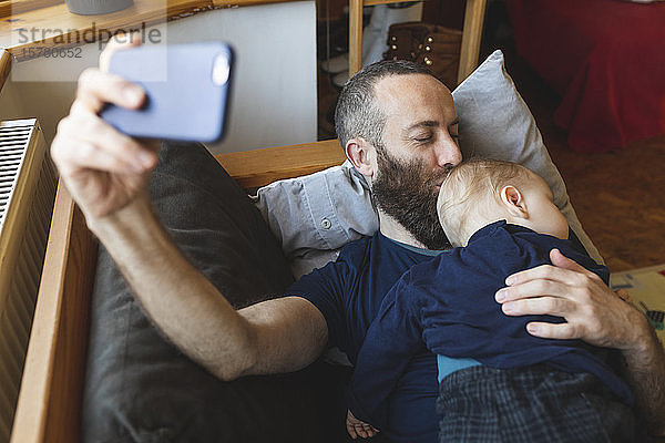 Mann  der sich mit seinem niedlichen schlafenden Sohn auf dem Sofa ein Selfie nimmt