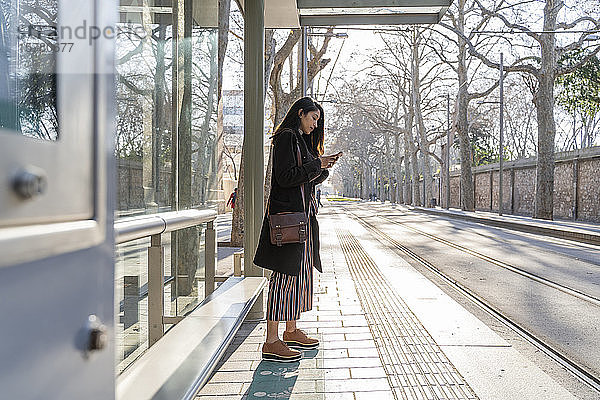 Junge Frau mit Smartphone wartet an der Straßenbahnhaltestelle