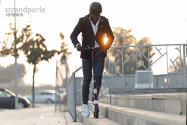 Junger Geschäftsmann mit Rucksack und Fahrradhelm fährt Roller bei Sonnenuntergang