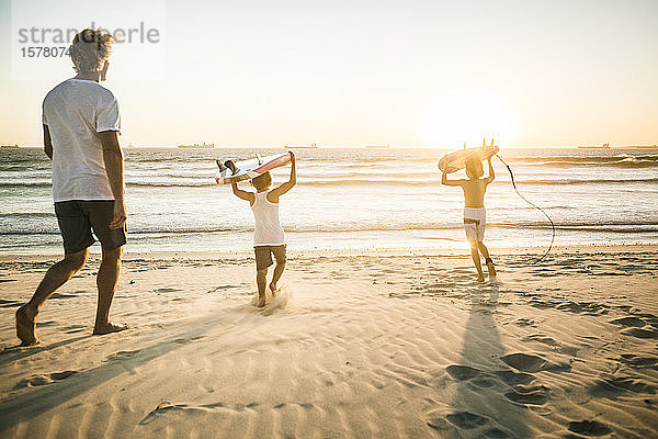 Vater und zwei Söhne mit Surfbrettern bei Sonnenuntergang am Strand spazieren