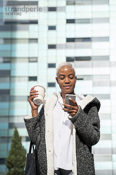 Stilvolle Businessfrau  die in der Stadt pendelt  Kaffee trägt und ein Smartphone benutzt