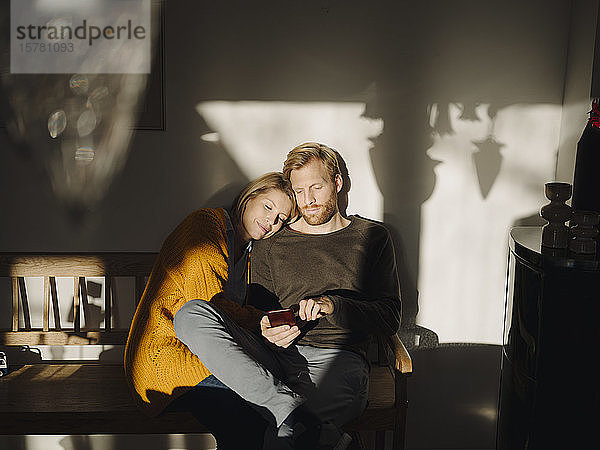 Entspanntes Paar sitzt zu Hause mit Mann auf Bank im Sonnenlicht und telefoniert