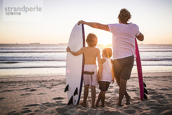 Rückansicht von Vater und Söhnen mit Surfbrettern  die bei Sonnenuntergang am Strand stehen