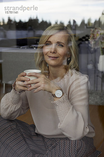 Lächelnde blonde Frau hält zu Hause Kaffeetasse hinter Fensterscheibe