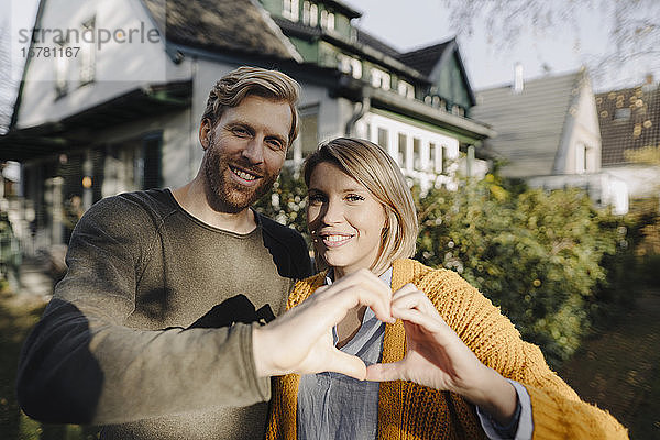 Porträt eines lächelnden Paares  das vor seinem Haus steht und mit den Händen ein Herz formt