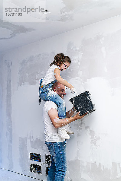 Mädchen sitzt auf den Schultern des Vaters und streicht die Wand ihres neuen Hauses