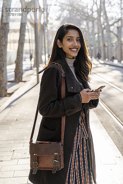 Porträt einer lächelnden jungen Frau mit Smartphone an der Straßenbahnhaltestelle