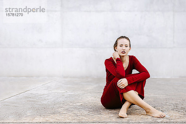 Porträt einer barfüssigen jungen Frau in rotem Kleid  die im Freien auf dem Boden sitzt