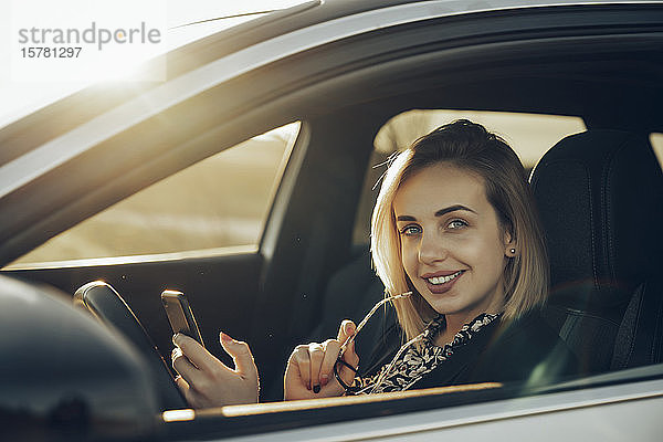 Porträt einer lächelnden jungen Frau mit Smartphone  die abends in ihrem Auto sitzt