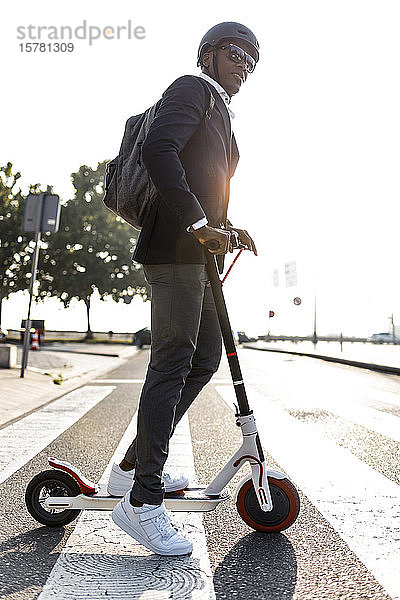 Geschäftsmann mit Rucksack und Fahrradhelm überquert bei Sonnenuntergang die Straße auf einem Schubsroller