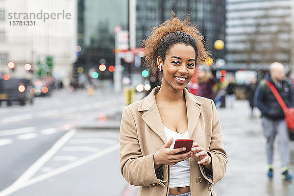 Lächelnde junge Frau mit Handy und Ohrstöpseln in der Stadt  London  Großbritannien