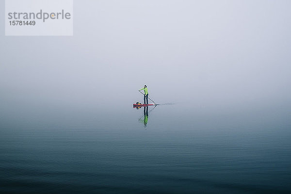 Mann steht beim Paddel-Surfen auf einem See im Nebel auf