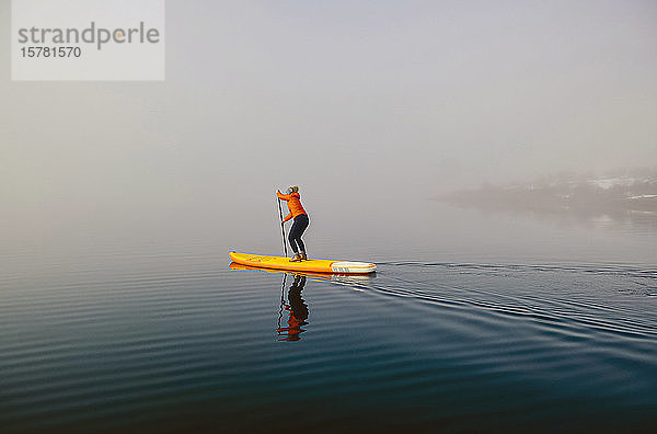 Frau steht beim Paddel-Surfen auf einem See im Nebel auf